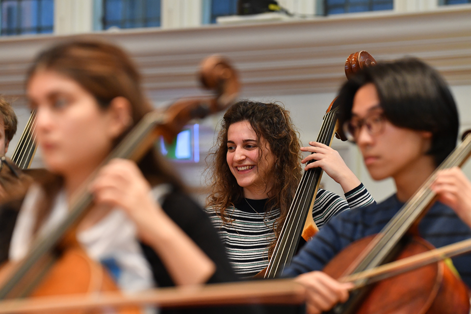 A female student with curly hair, holding a double bass in her left hand, smiling, while sitting in an orchestra rehearsal.